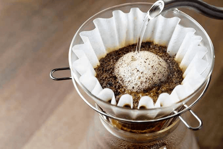 hot water is poured over coffee grounds in the top of a Chemex coffee brewer