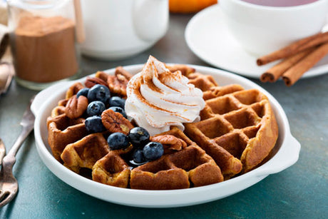 A round Belgian waffle topped with blueberries, pecans, and a dollop of whipped cream, sprinkled with cinnamon